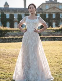 Model in a Allure Bridgerton style BR1007 wedding dress in front of a royal looking english countryside house made of red brick with white window and door frames but no white columns. 